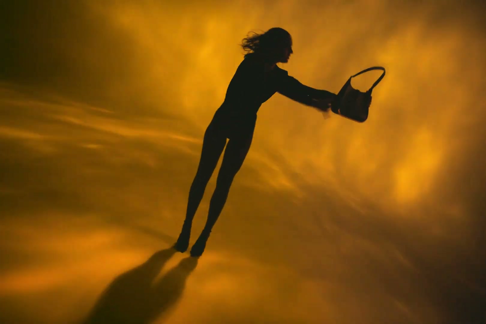 Silhouetted figure reaching forward with a handbag against a golden backdrop — still from the Vaulie ICON campaign film, scored by Florian Knapp